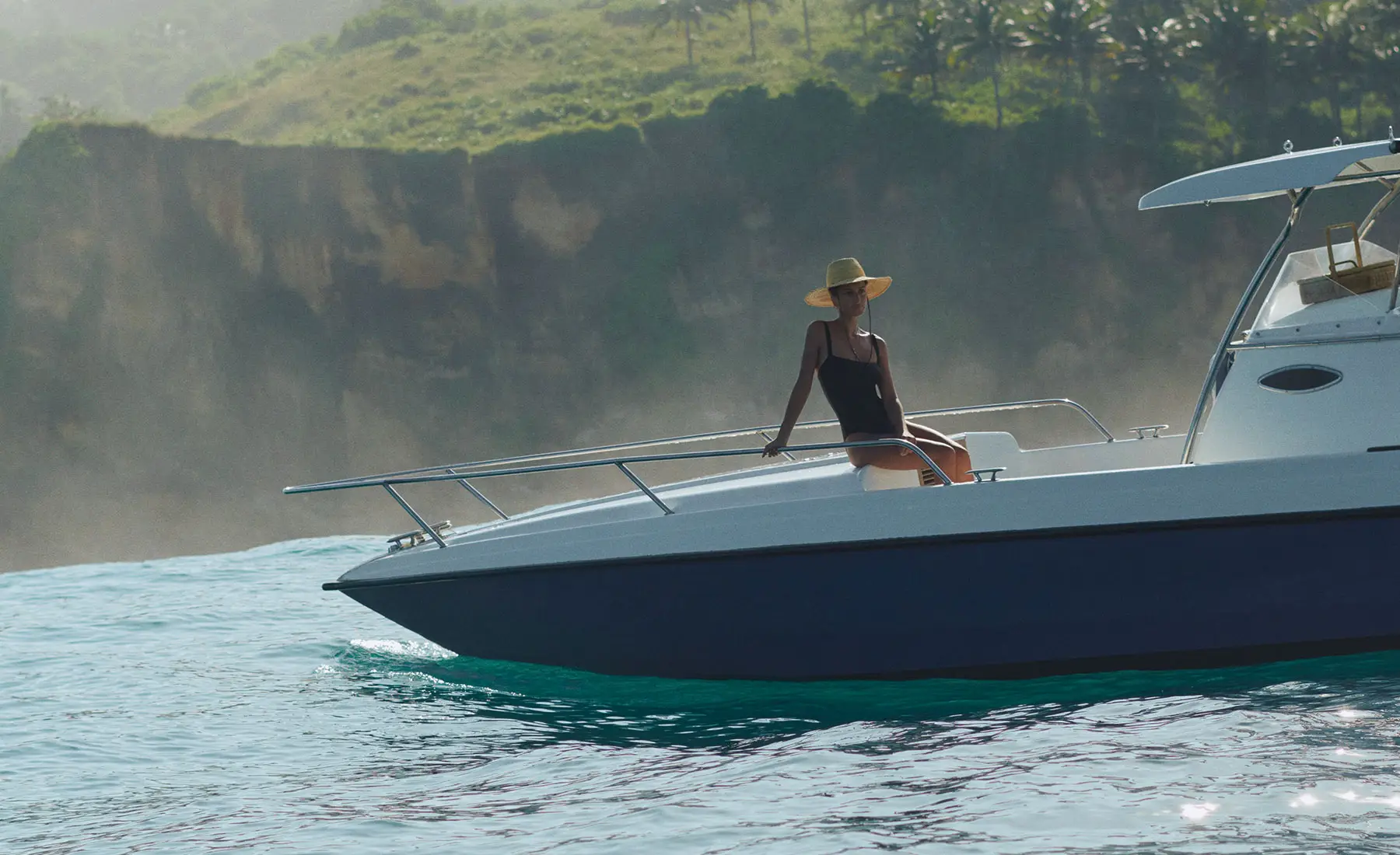 A woman in a sunhat and black swimsuit relaxes at the bow of a luxury speedboat off the coast of Sumba, with dramatic cliffs and lush greenery in the background—capturing the spirit of ocean exploration at NIHI Sumba.