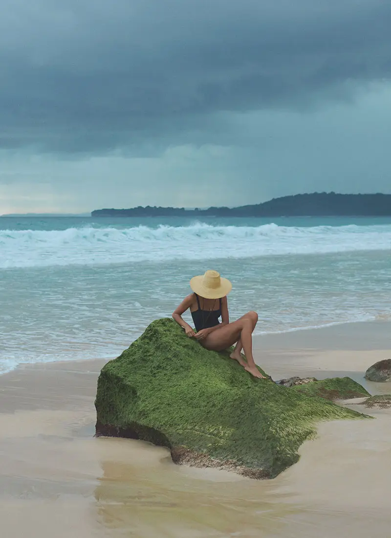 Woman in a black swimsuit and sunhat relaxing on a moss-covered rock at Nihiwatu Beach, NIHI Sumba, under dramatic cloudy skies.