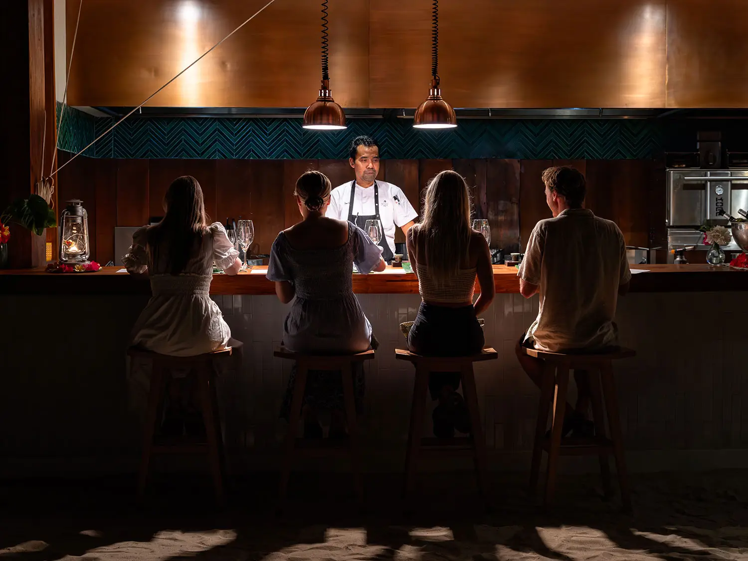 A group of guests seated at the Chef’s Table at Nio, immersed in the intimate dining experience as the chef stands behind the counter. The setting is enhanced by warm lighting and elegant decor, reflecting NIHI Sumba’s signature fusion of East and West cuisines.