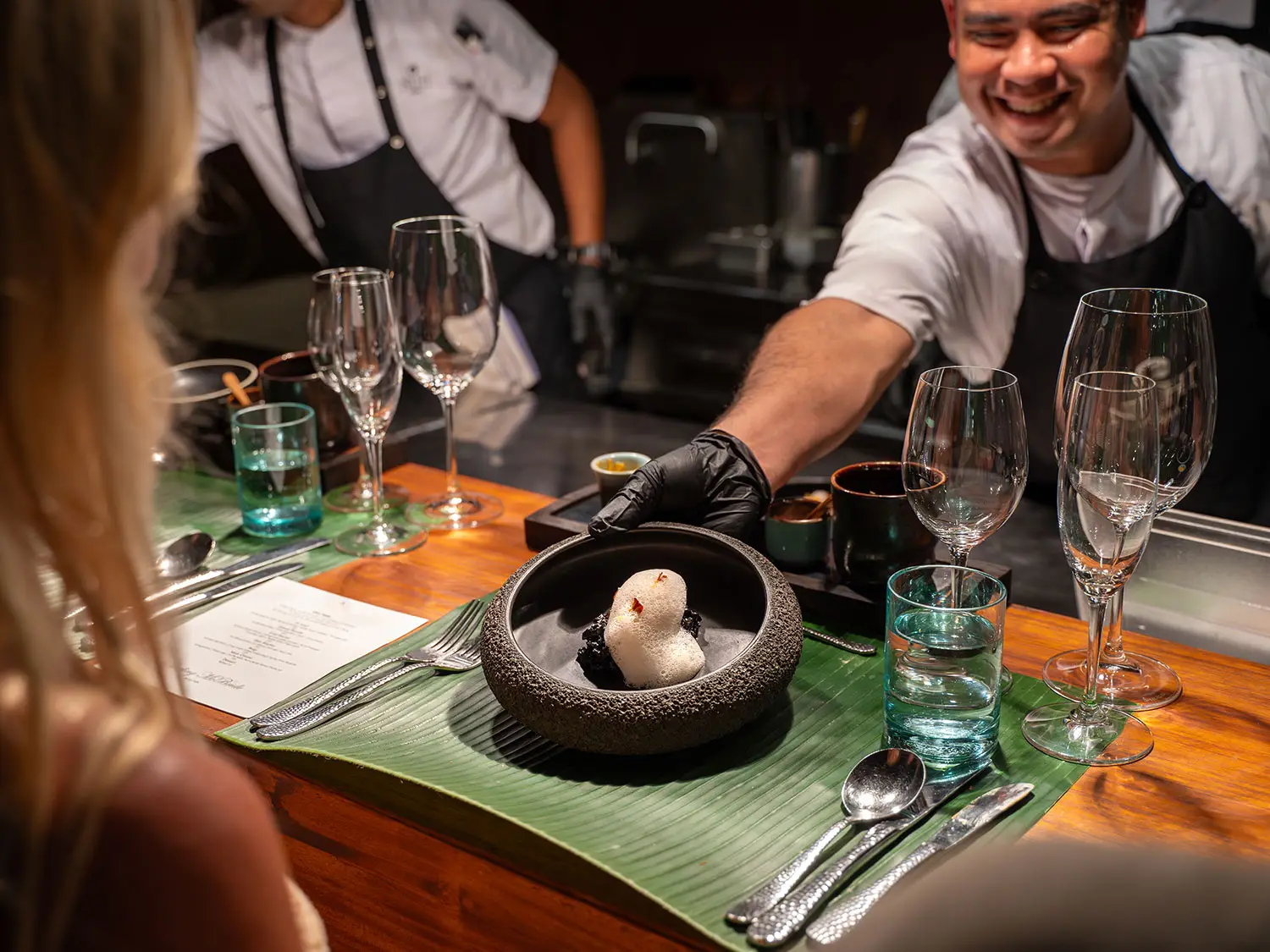 A chef at the Chef's Table at Nio presenting a carefully crafted dish in a stone bowl, showcasing the fusion of East and West culinary traditions, while guests enjoy an intimate and personalized dining experience.
