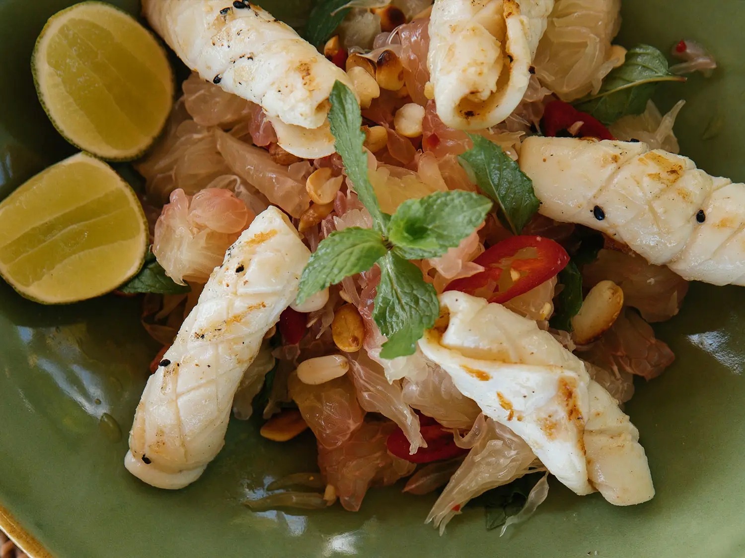 A vibrant and fresh bowl of grilled seafood salad with mint, chili, and pomelo, garnished with lime wedges and peanuts, offering a delightful Indonesian dish at NIHI Sumba.