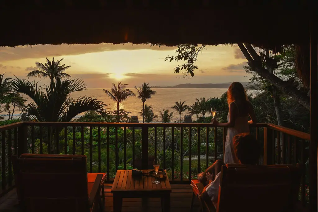 Guests enjoying a sunset view over the Indian Ocean from the private balcony of Kanatar villa at NIHI Sumba, surrounded by tropical palms and tranquil luxury.