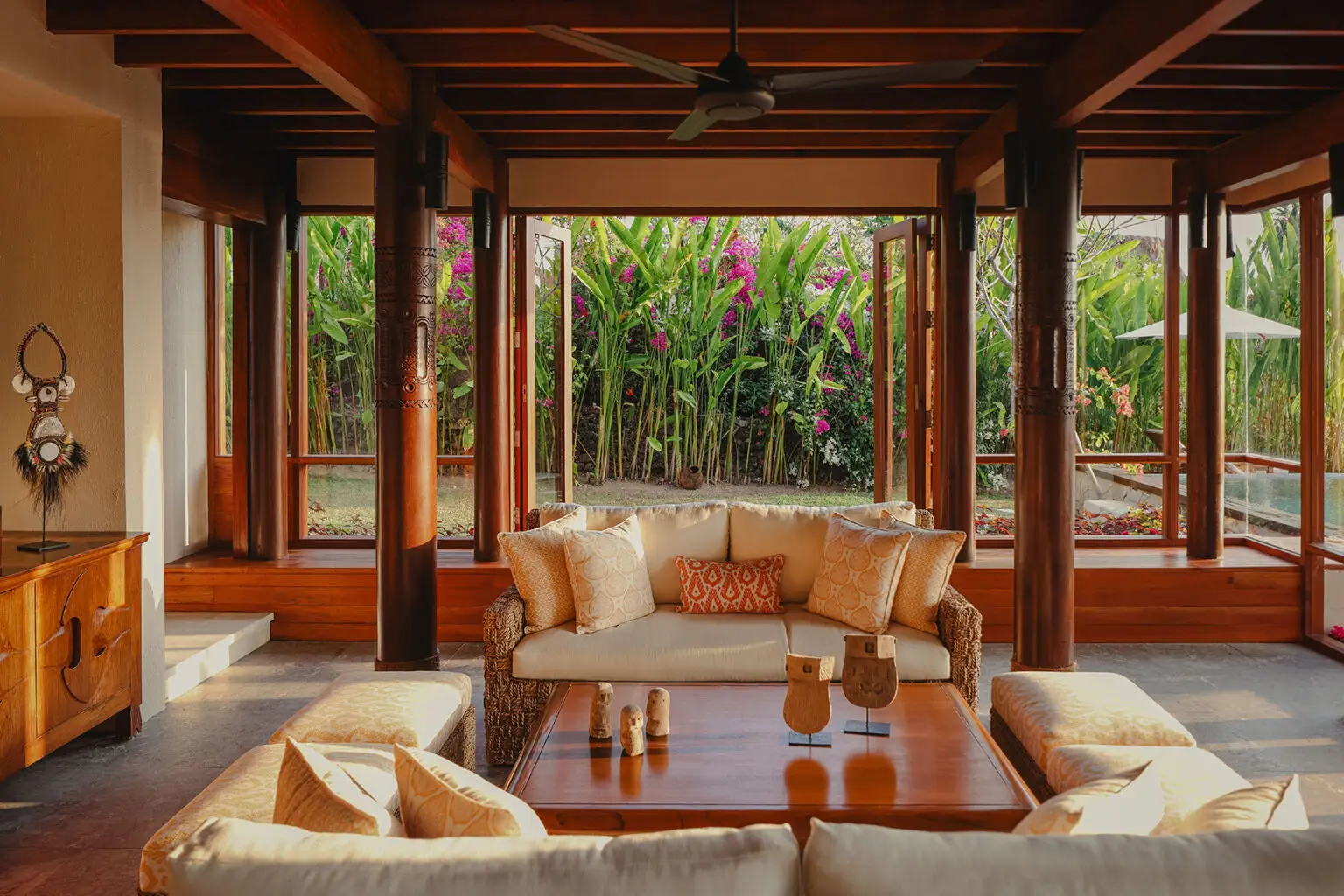 Sunlit indoor lounge at Kanatar villa in NIHI Sumba, featuring a cozy rattan sofa set with views of lush tropical foliage and blooming flowers just beyond the open windows.