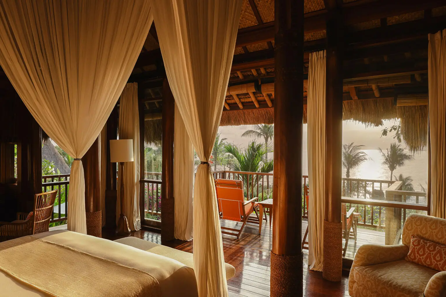 Elegant canopy bed in a sunlit bedroom at NIHI Sumba’s Kanatar villa, featuring rich wooden interiors, flowing curtains, and a private balcony overlooking tropical palm trees and an ocean view.