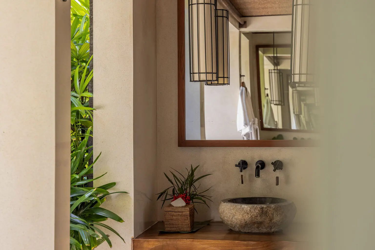 The bathroom area of the Kanatar Villa at NIHI® Sumba, featuring a stone sink, dark metal faucets, a large mirror, and tropical greenery visible outside the room.