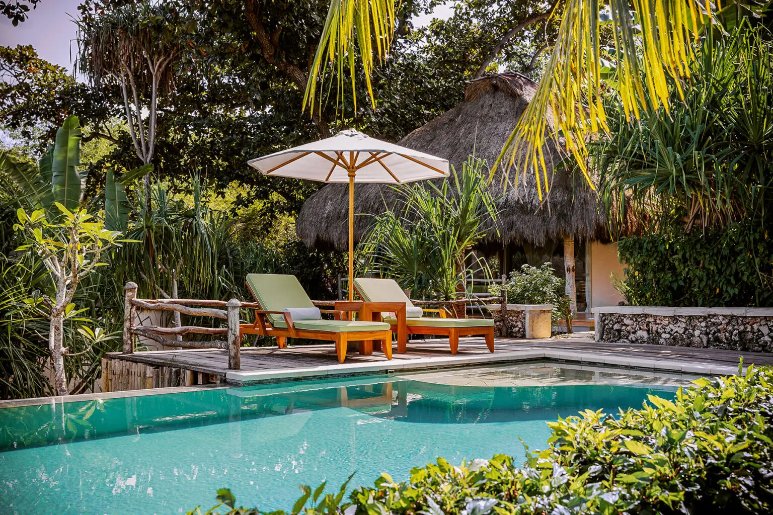 Cozy poolside setting at the Kasambi 1-Bedroom Villa, NIHI Sumba, featuring sun loungers beneath a white umbrella and a thatched-roof villa surrounded by lush tropical gardens near Occy’s Left.