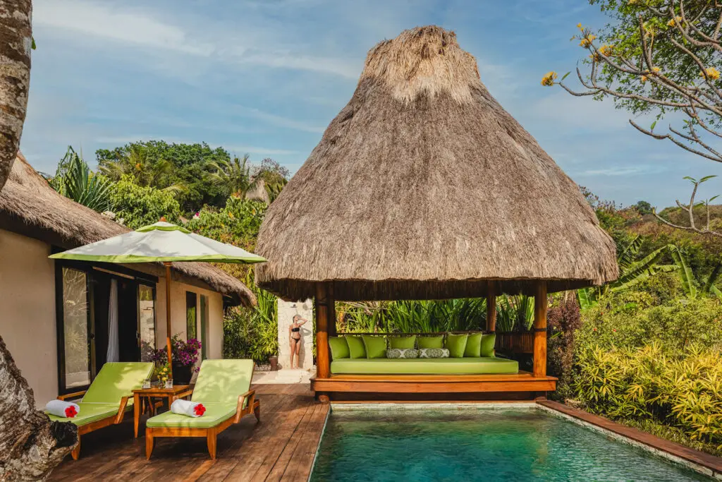 Private plunge pool and shaded bale at Lulu Amahu Saudara, a two-bedroom villa at NIHI® Sumba designed for families, surrounded by lush gardens and furnished with green sun loungers and cushions.