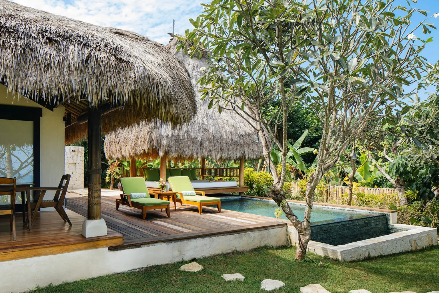 A serene outdoor lounge area at the Lulu Amahu Saudara Villa, with two comfortable sun loungers and a large parasol by the private plunge pool. The stylish wooden deck features a cozy seating area with green cushions, providing a perfect setting to relax and enjoy the lush tropical surroundings. The vibrant greenery and tropical plants complement the peaceful atmosphere of this luxurious villa at NIHI Sumba.