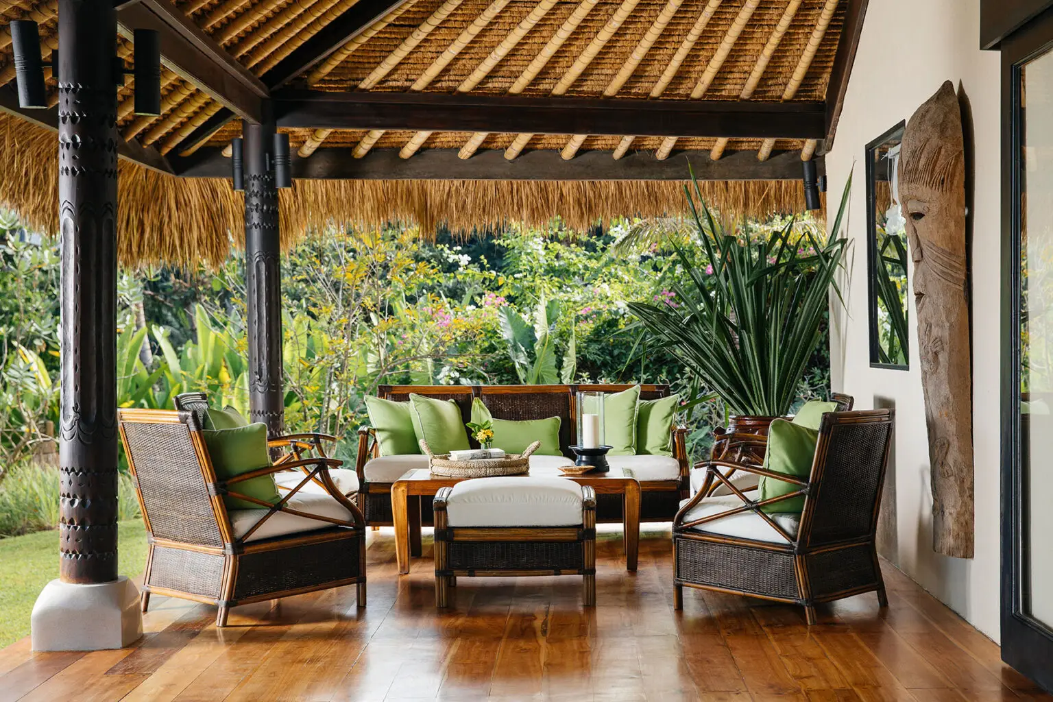The outdoor lounge area of the Lulu Amahu Saudara Villa at NIHI Sumba. The space is adorned with stylish rattan furniture, featuring a sofa with soft cushions in earthy tones of green. An intricately designed wooden coffee table sits in the center, and the surrounding walls are decorated with traditional artwork. The open-air environment is enhanced by a thatched roof and lush tropical plants visible in the background, creating a serene, inviting atmosphere for relaxation.