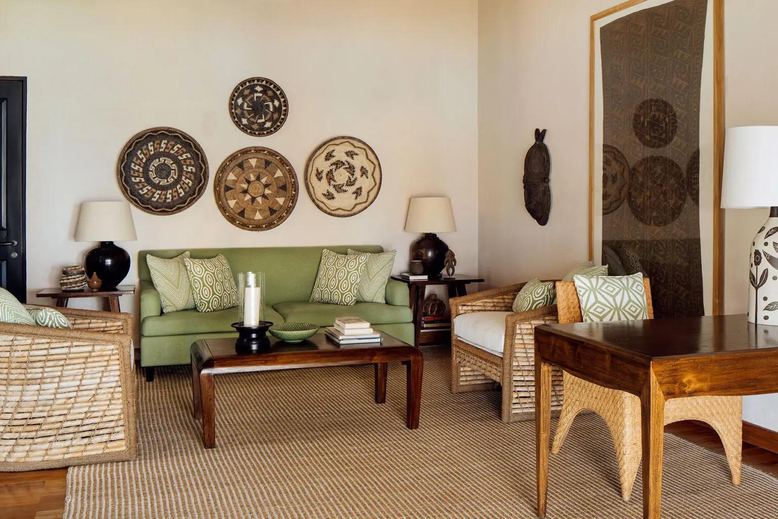 Interior view of the Lulu Amahu Saudara Villa at NIHI Sumba, showcasing a stylish living area. The space features a green sofa adorned with patterned cushions, wicker armchairs, and a wooden coffee table. The walls are decorated with traditional woven circular art pieces, creating a warm, inviting atmosphere with a blend of tropical and cultural elements.