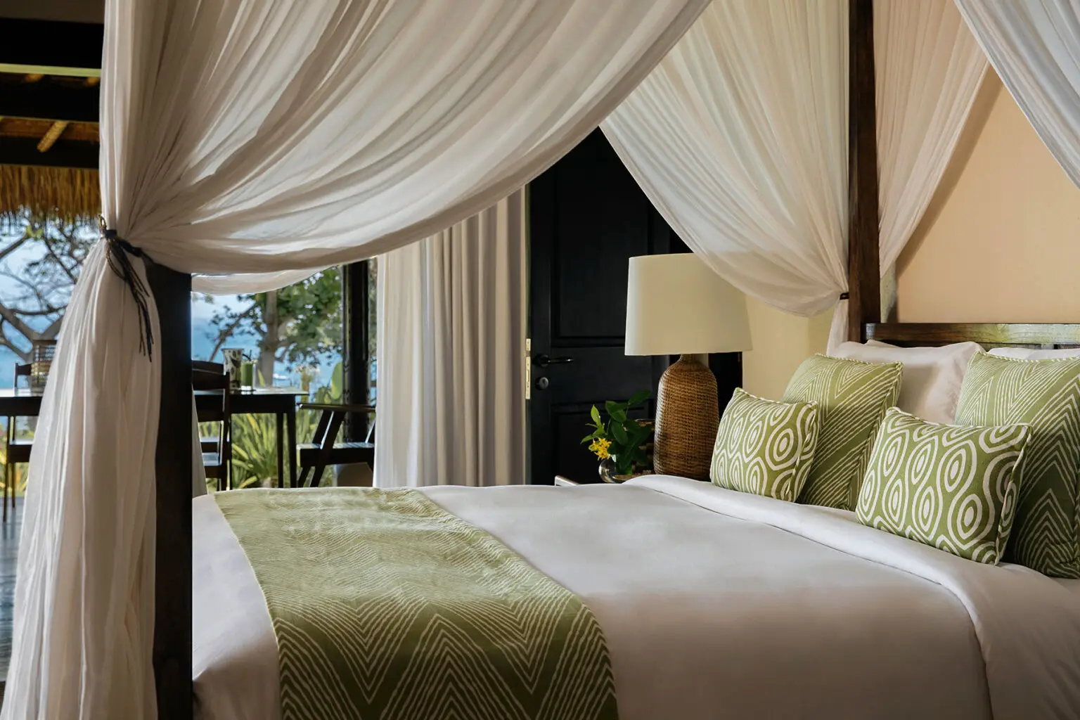Master bedroom at Lulu Amahu Saudara Villa, NIHI® Sumba, featuring a bed framed by flowing white drapery, green and patterned cushions, a rustic wooden lamp, and a window overlooking lush tropical greenery in a serene and luxurious setting.
