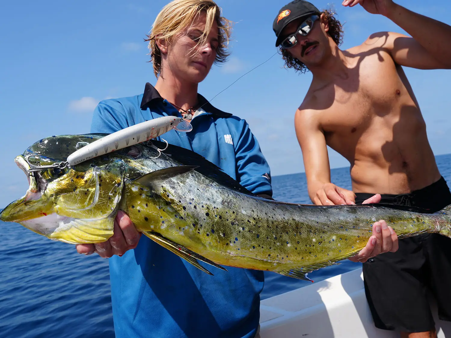 Two anglers proudly display their catch of a large mahi-mahi fish aboard a fishing boat near NIHI Sumba. One angler holds the fish, while the other stands beside him on the boat under a clear blue sky, showcasing their successful fishing adventure.