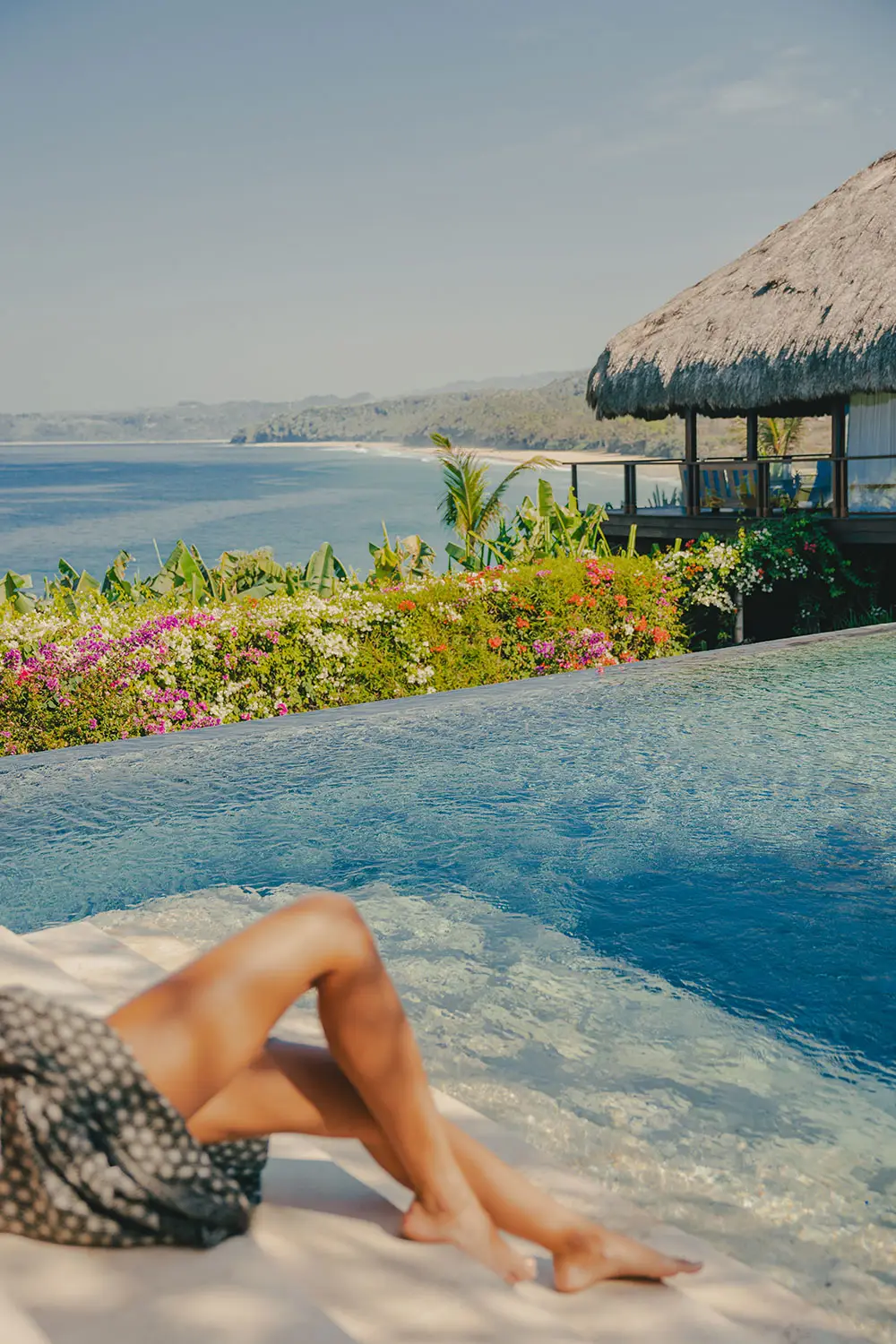 A guest relaxing in Raja Mandaka’s infinity pool overlooking the ocean, surrounded by vibrant tropical flowers and coastal hills under a clear blue sky.