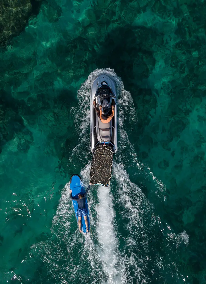 An aerial view of a jet ski towing a surfer over clear turquoise waters at NIHI® Sumba, showcasing the resort's water sports experiences in the Indian Ocean.