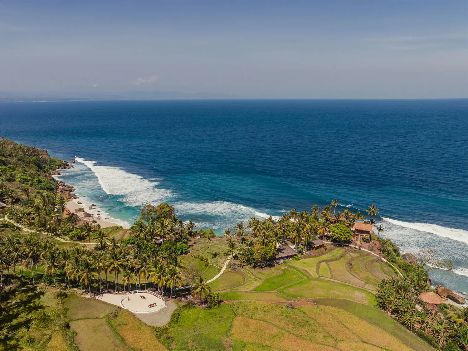 Aerial view of Nihioka Spa at NIHI® Sumba, showing its coastal cliffside location surrounded by terraced rice fields, tropical jungle, and a nearby horse ring, with sweeping views of the Indian Ocean below.