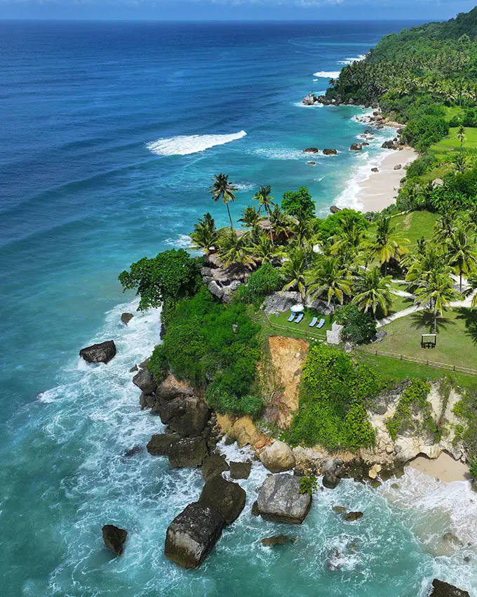 Aerial view of Nihioka Spa, nestled along the turquoise coastline of Sumba and surrounded by lush green fields and swaying palm trees—capturing the serene and secluded cliffside setting.