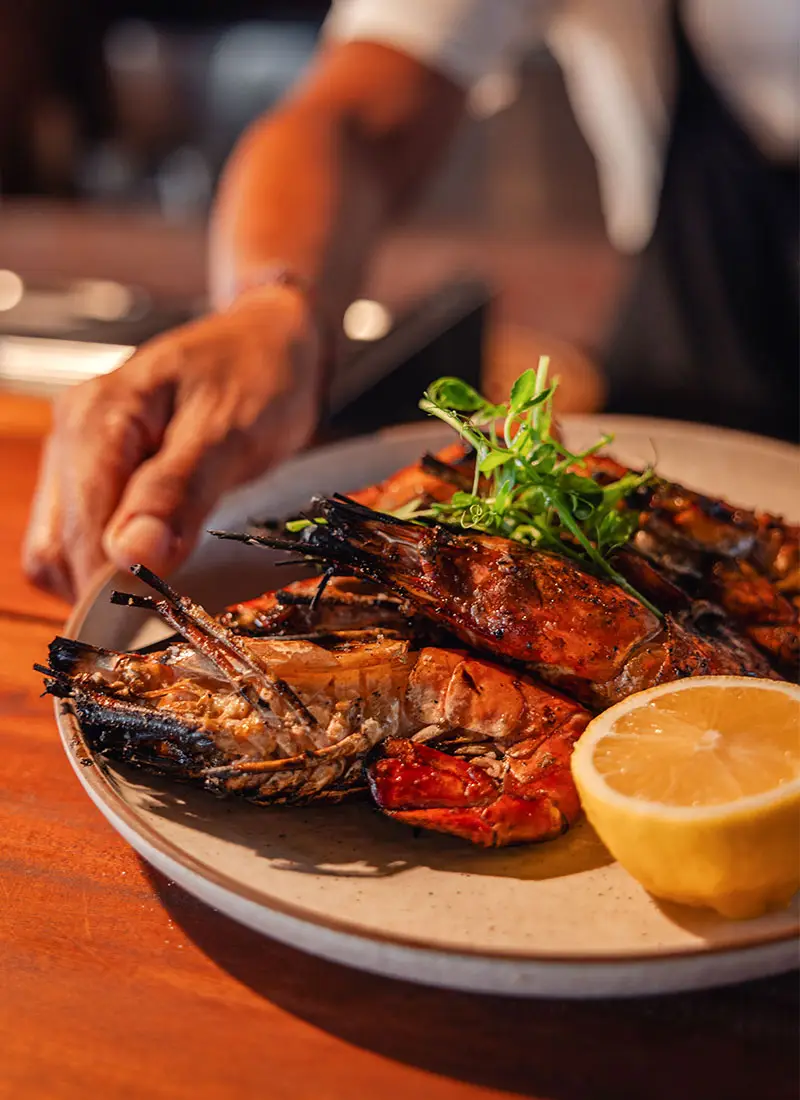 Close-up of grilled prawns garnished with fresh herbs and a slice of lemon, served as part of the seafood feast at Ombak Restaurant, NIHI Sumba. The dish showcases the fresh, locally sourced ingredients that are central to the resort's culinary offerings.