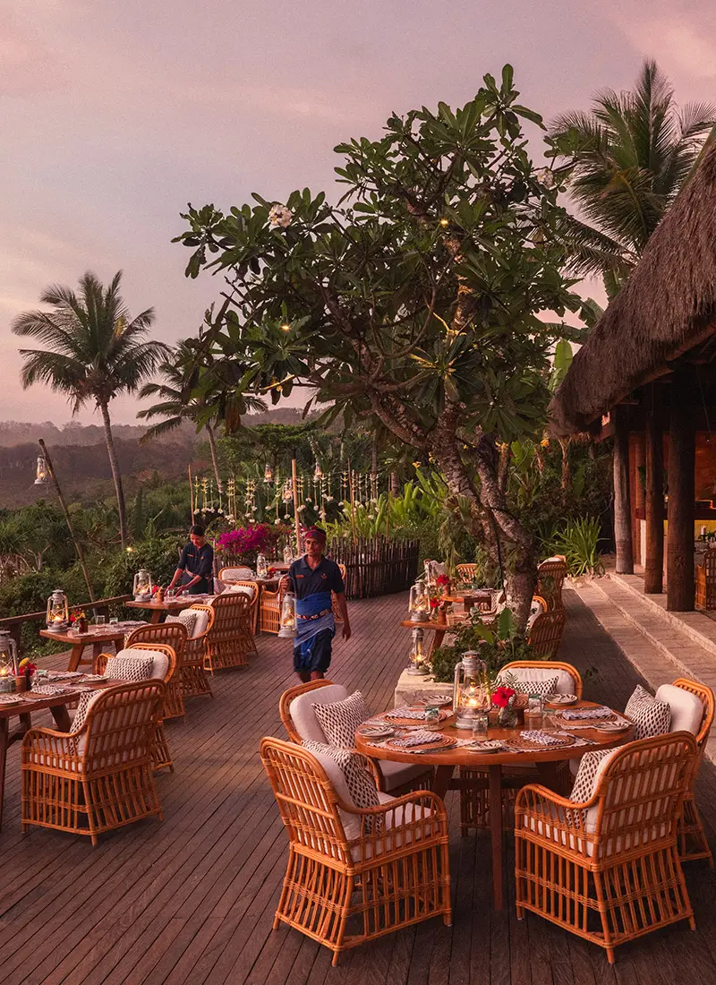 Outdoor dining setup at Ombak Restaurant, NIHI Sumba, during sunset, with elegant wicker tables set with plates and candles, surrounded by lush greenery and overlooking the 2.5km-long Nihiwatu Beach, as staff prepare the area to create a tranquil and romantic atmosphere.
