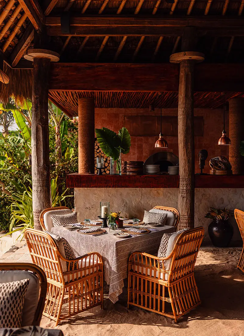 A beautifully set table at Ombak Restaurant, NIHI Sumba, featuring wicker chairs, a linen tablecloth, and tropical decor. The open-air dining space is surrounded by lush greenery, offering a serene and intimate setting for guests to enjoy fresh, locally sourced meals.