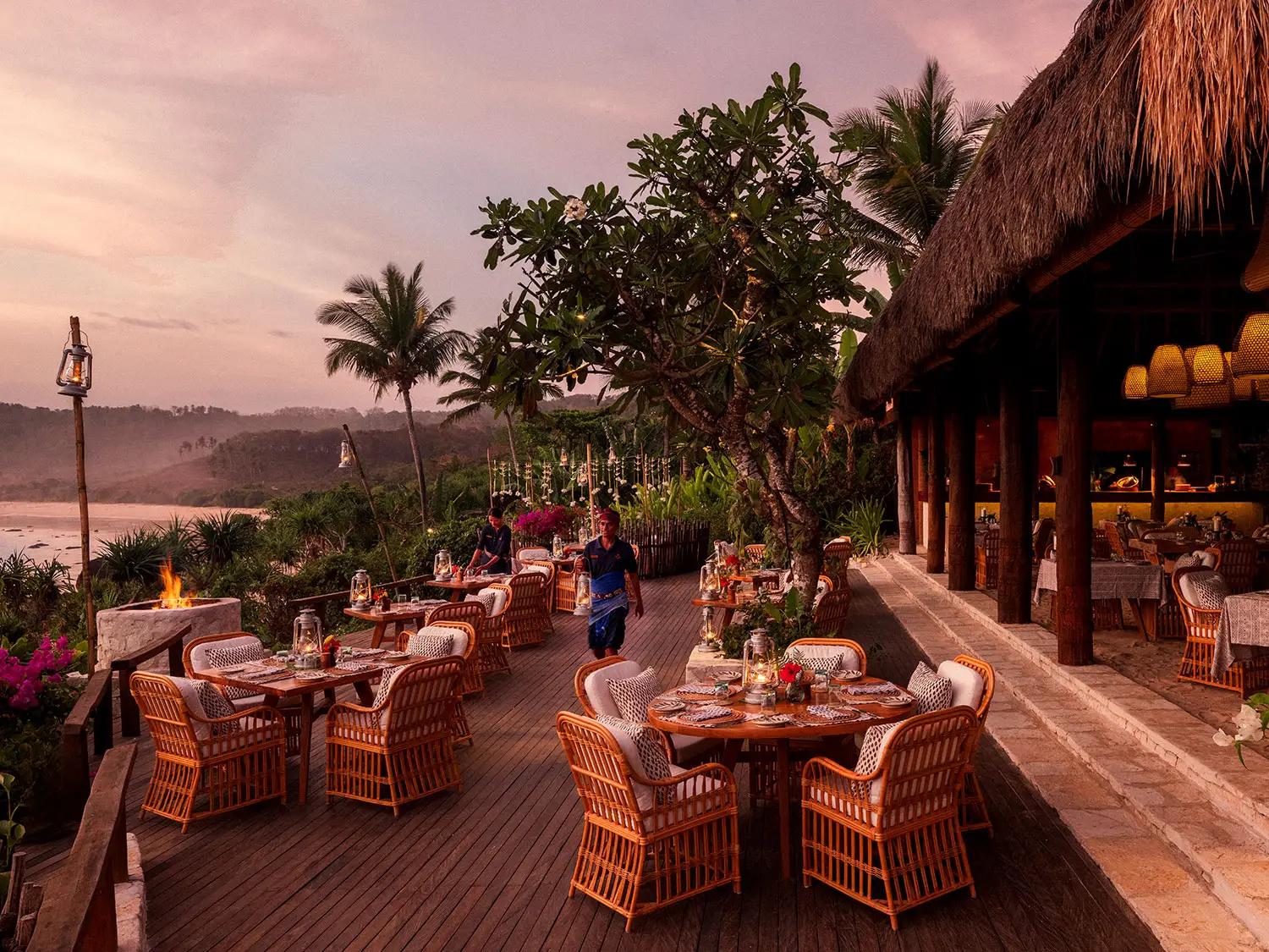 Outdoor dining setup at Ombak Restaurant, NIHI Sumba, during sunset, with elegant wicker tables set with plates and candles, surrounded by lush greenery and overlooking the 2.5km-long Nihiwatu Beach, as staff prepare the area to create a tranquil and romantic atmosphere.