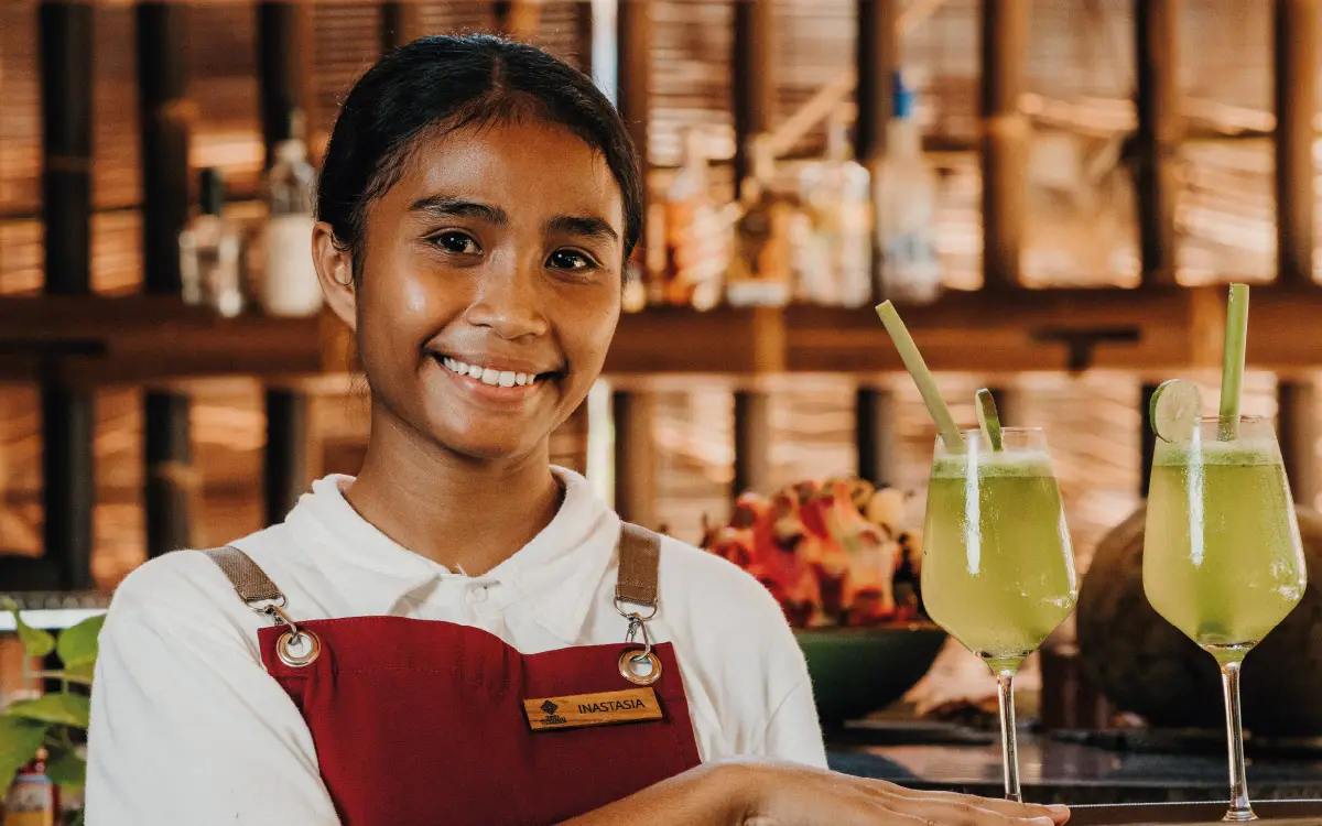 A smiling student from the Sumba Hospitality School serves iced tea at a bamboo training bar, demonstrating practical hospitality skills in a sustainable learning environment.