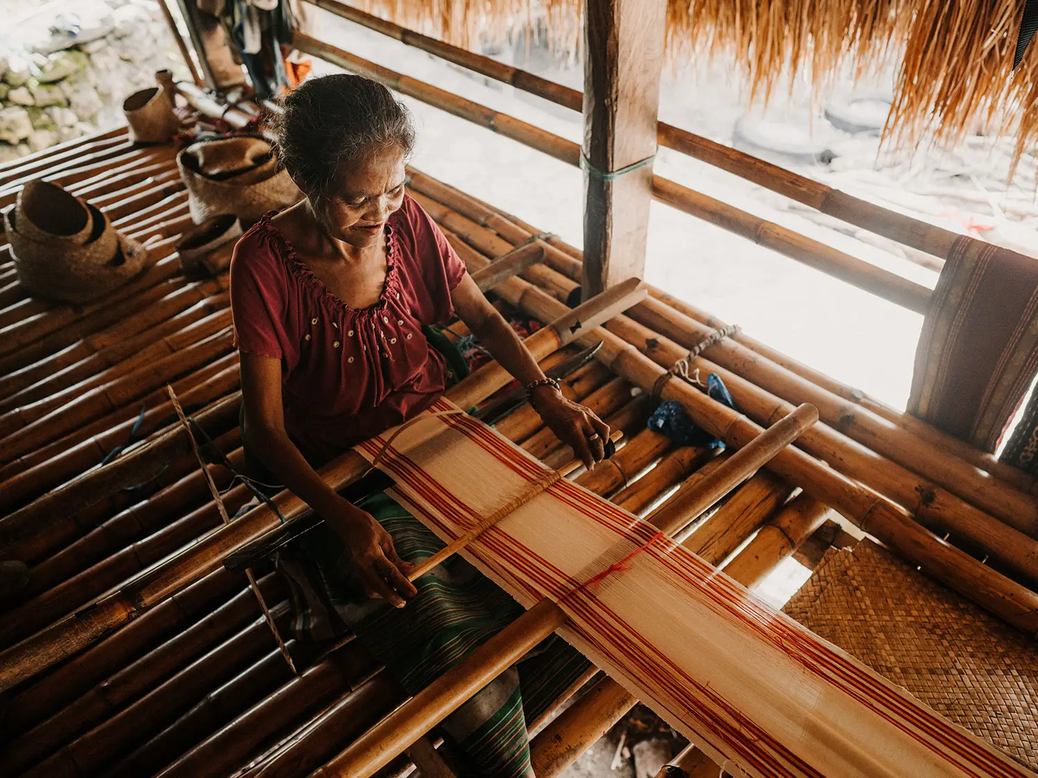 A Sumbanese artisan seated at a traditional wooden loom, weaving a vibrant tenun ikat textile at NIHI Sumba, surrounded by colorful handwoven fabrics that reflect generations of cultural craftsmanship.