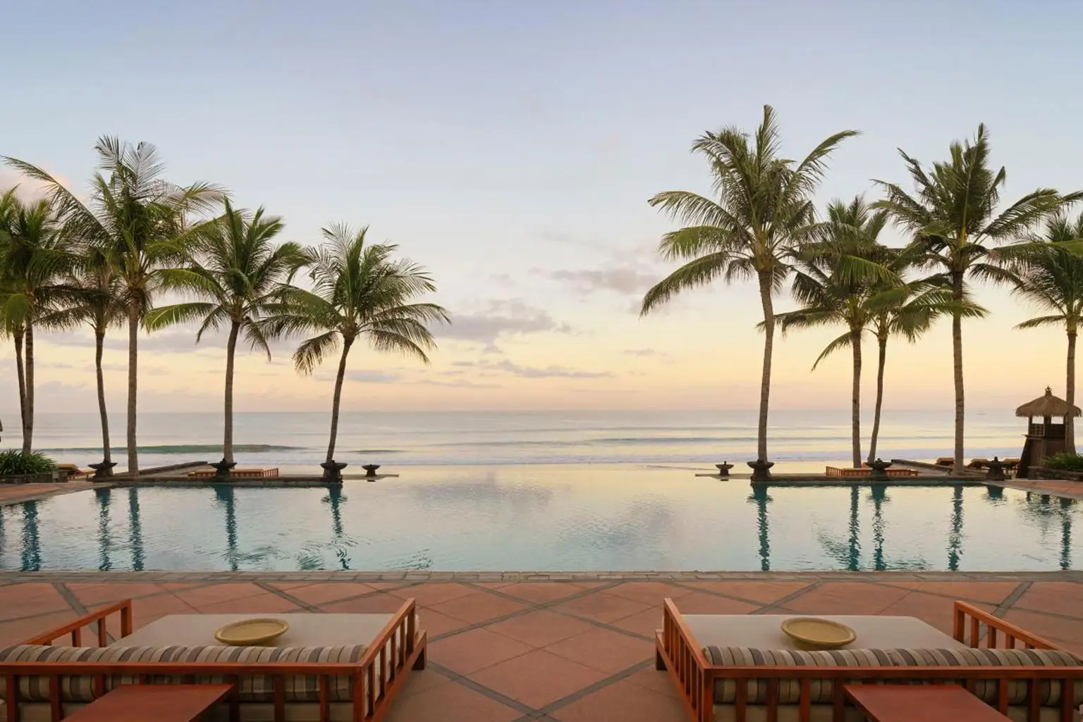 A view of the stunning infinity pool at The Legian Seminyak, Bali, with the Indian Ocean stretching into the distance. Surrounded by lush palm trees, the pool area offers guests a serene and picturesque setting, perfect for a luxurious island getaway.