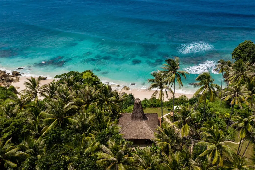 A stunning aerial view of Villa Rahasia, located within the Nihioka Spa at NIHI Sumba, nestled amidst lush tropical greenery and overlooking the tranquil Indian Ocean. This secluded villa offers ultimate privacy with direct access to a pristine beach.