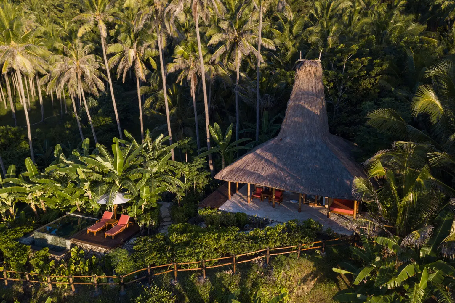 Aerial view of Villa Rahasia, a secluded hideaway within the Nihioka Spa at NIHI Sumba, surrounded by lush tropical greenery and tall palm trees. The villa features a private plunge pool beside a wooden deck with two sun loungers under a thatched roof, creating an idyllic space for relaxation.