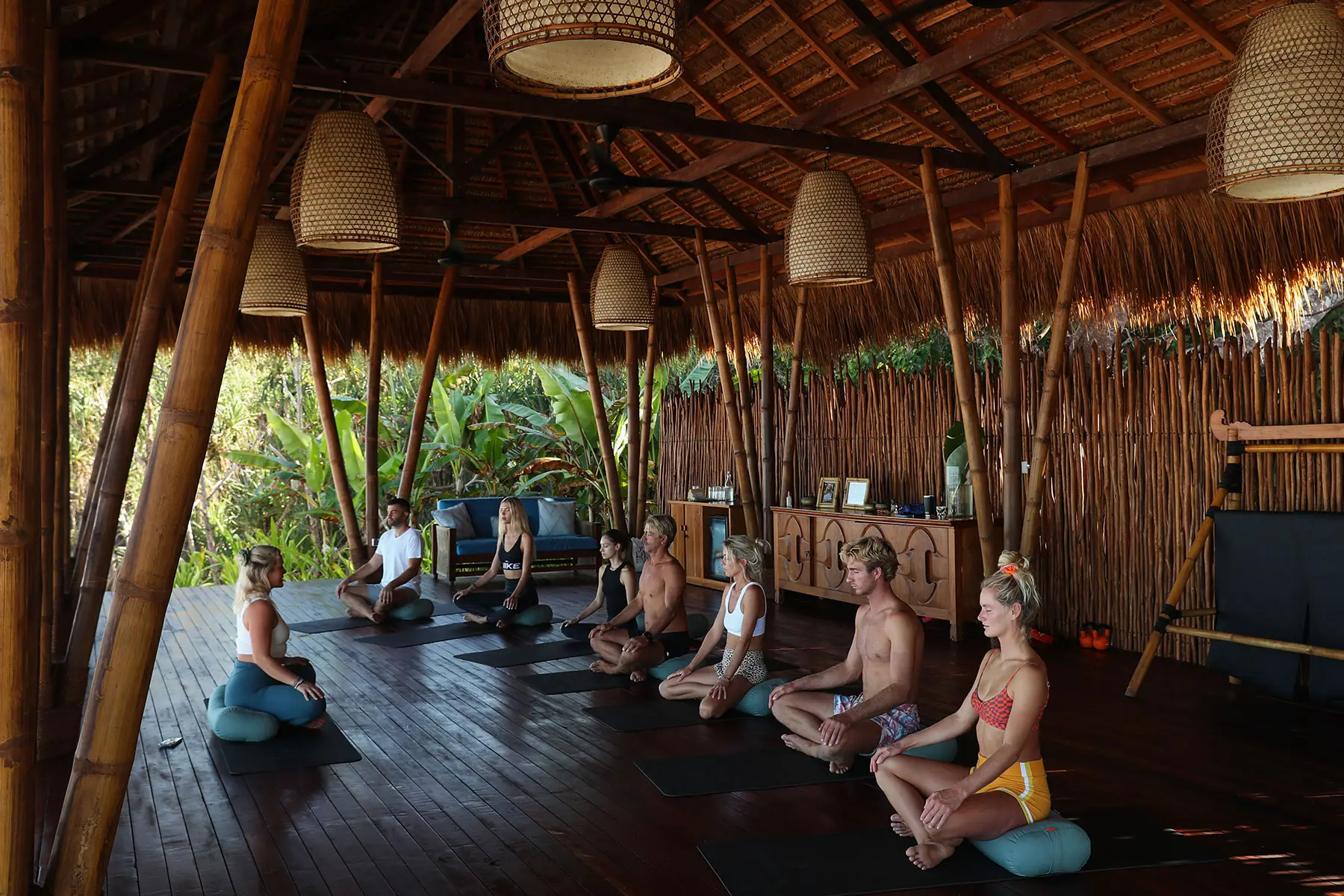 A group of guests practicing yoga in the Yoga Pavilion at NIHI Sumba, with bamboo structures surrounding them and lush greenery visible outside, as part of the Wild Wellness program.
