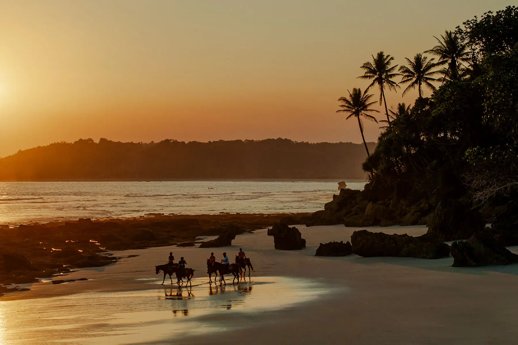 Guests on horseback riding along the shore at sunset during a festive Easter celebration on Nihiwatu’s 2.5 km beach, with giant colorful eggs nestled in the sand beneath swaying palms and golden skies.