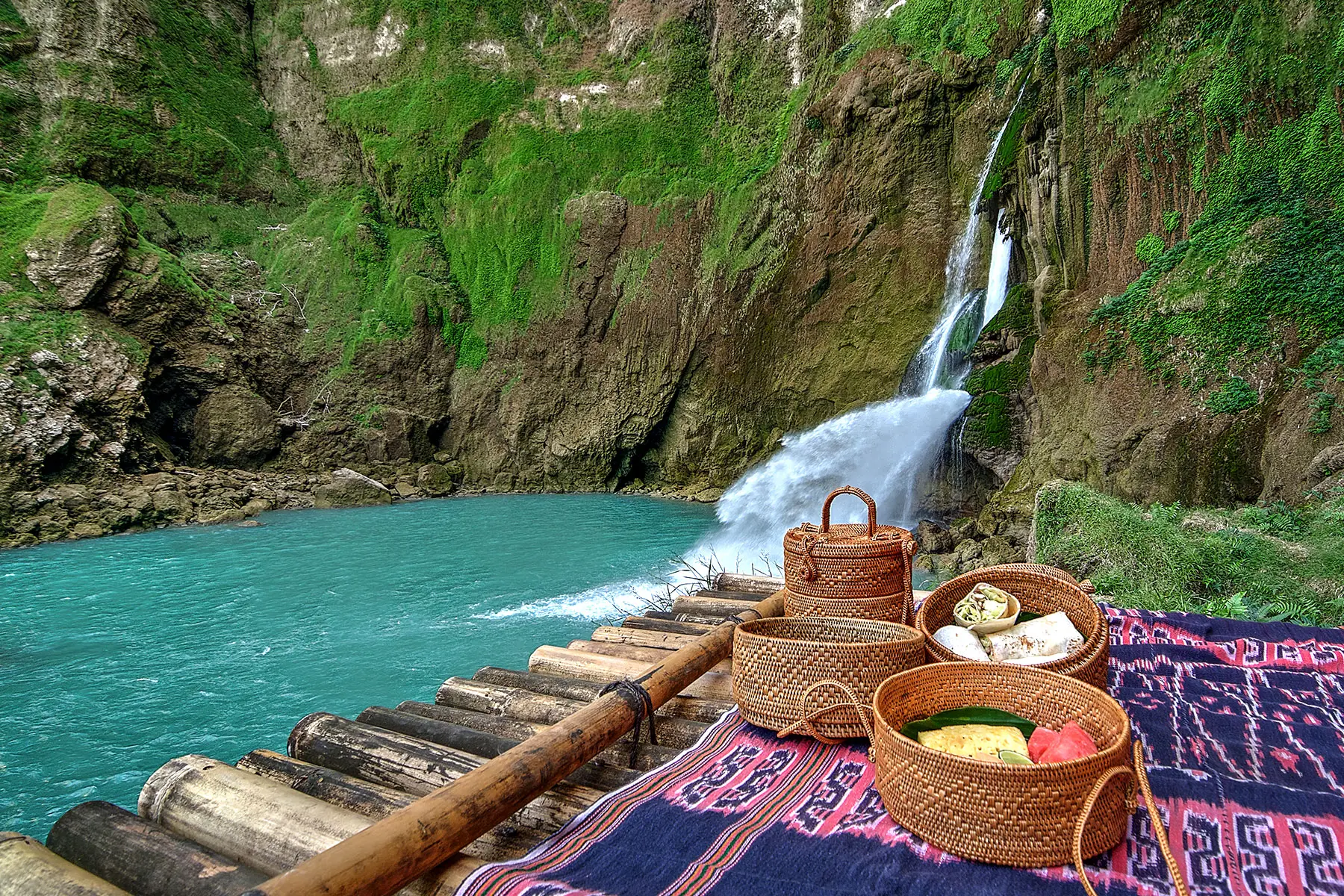 A scenic picnic setting beside the Blue Waterfall at NIHI Sumba, featuring traditional woven baskets filled with local delicacies placed on a bamboo platform and an ikat blanket, surrounded by lush greenery.