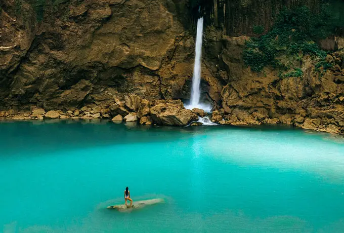 Traveler standing on a rock in turquoise blue lagoon beneath a stunning Matayangu Waterfall in Sumba Island, Indonesia.