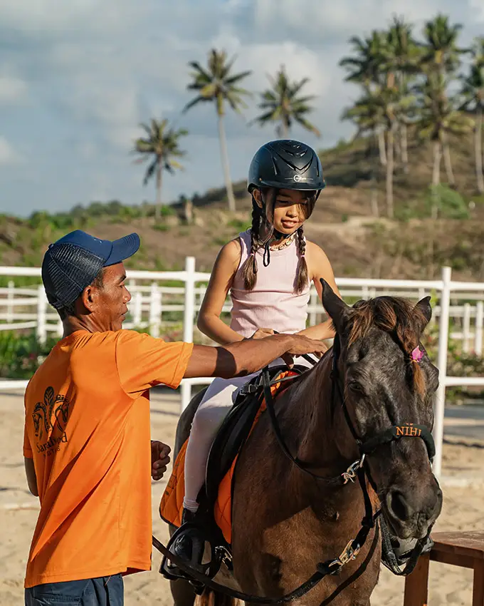 Sumba pony trainer guiding a young girl during a riding lesson at NIHI Sumba's Sandalwood Stables, with tropical scenery in the background.