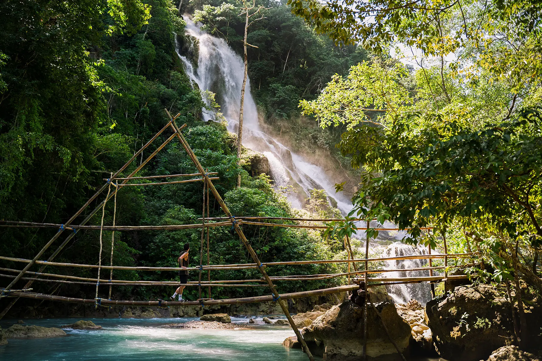 A guest crossing a bamboo suspension bridge in front of Lapopu Waterfall, surrounded by dense jungle and cascading tiers of water in the remote highlands of Sumba Island.
