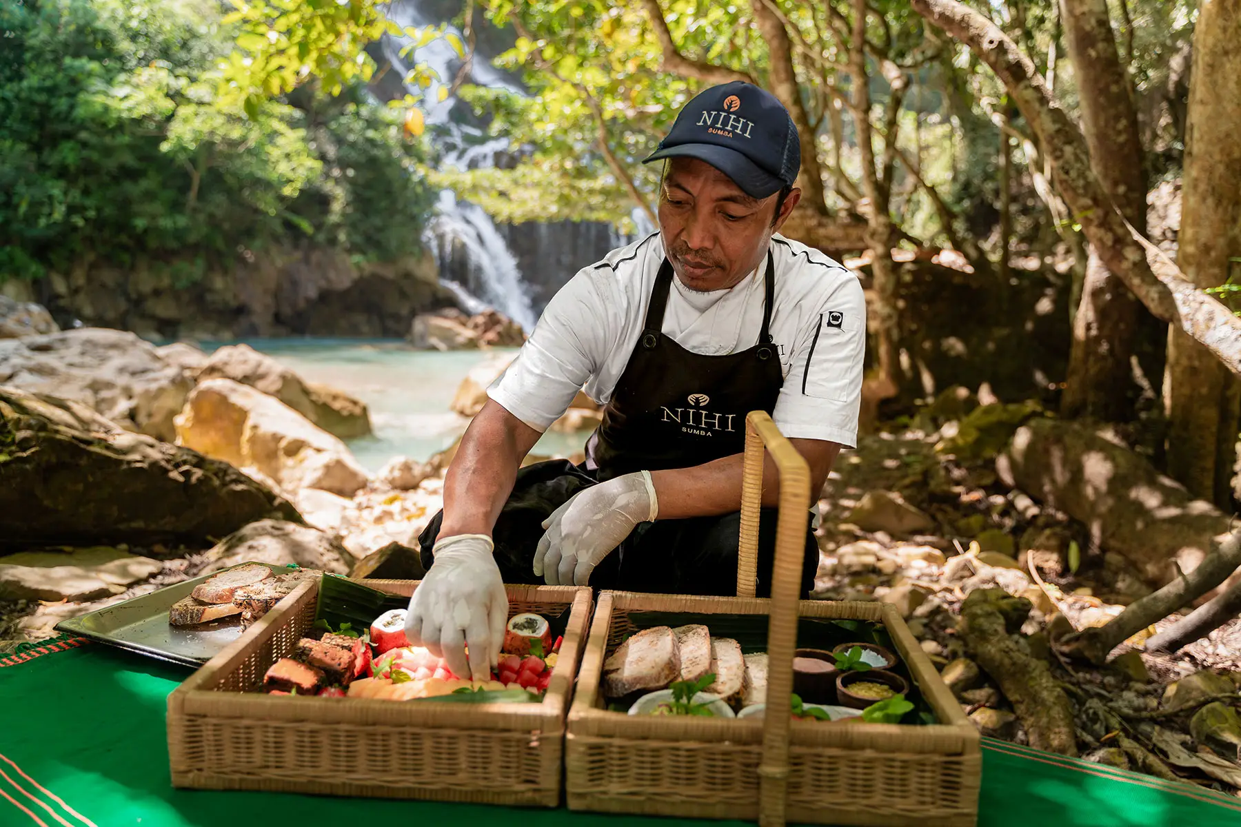A NIHI Sumba chef prepares a gourmet riverside picnic with fresh tropical fruits, artisanal bread, and local delicacies beside the serene Lapopu Waterfall in the Sumbanese jungle.