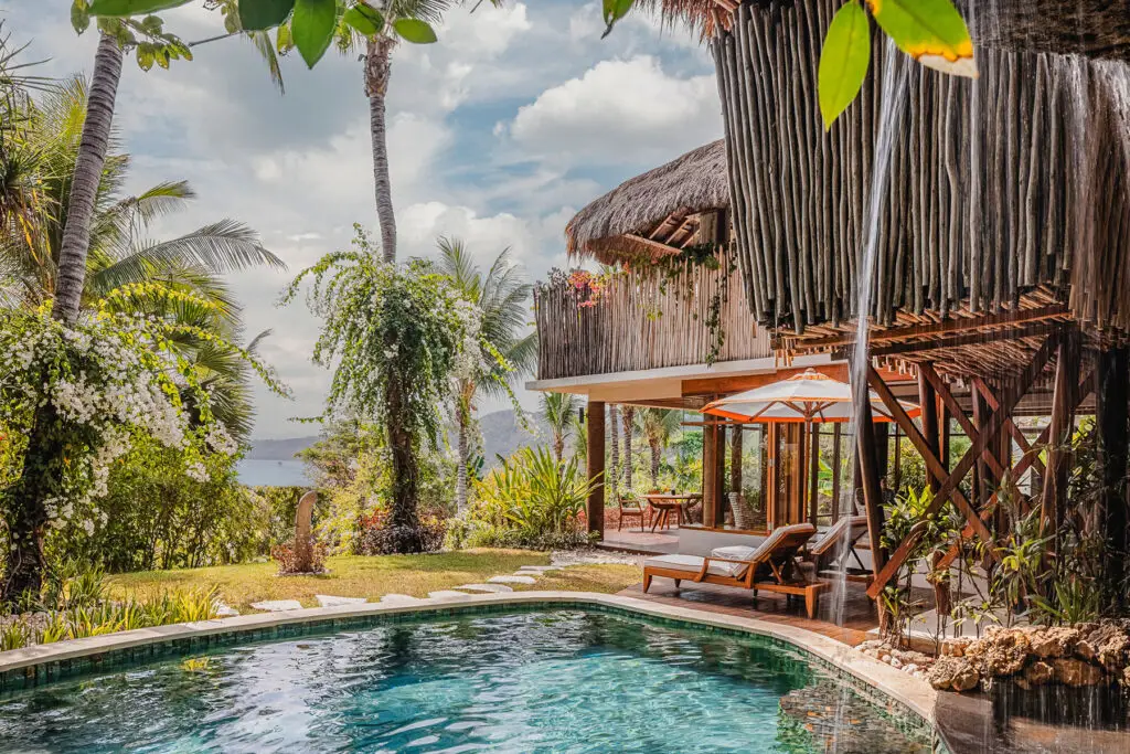 Private pool at Mandaka Sumba House with lush tropical gardens, palm trees, and traditional thatched architecture, offering breathtaking views of the ocean at NIHI Sumba
