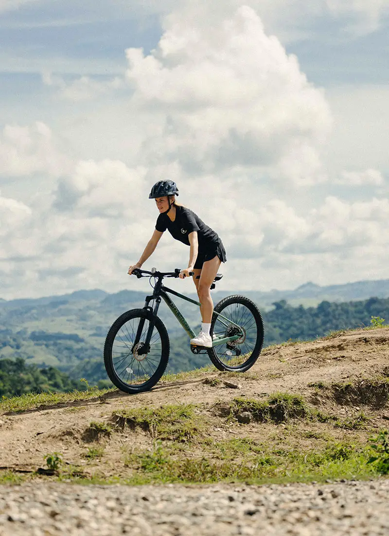 A woman rides a mountain bike along a dirt trail overlooking the lush highlands of Sumba, Indonesia, during a scenic cycling excursion organized by NIHI® Sumba.