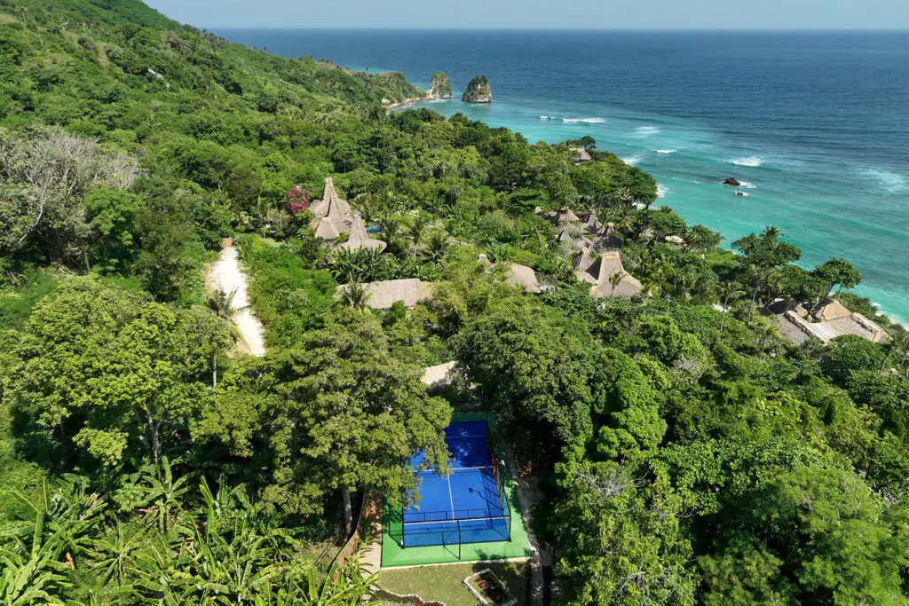 Aerial view of NIHI Sumba’s vibrant padel court set amid lush tropical gardens, with guests playing a match against the backdrop of thatched villas and the Indian Ocean — a highlight of the resort’s Active Wellness program.