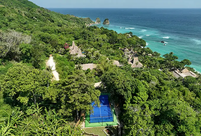 Aerial view of NIHI Sumba’s vibrant padel court set amid lush tropical gardens, with guests playing a match against the backdrop of thatched villas and the Indian Ocean — a highlight of the resort’s Active Wellness program.