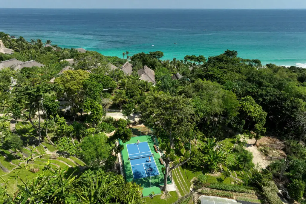 Aerial view of the Pickleball Court at NIHI Sumba, surrounded by lush jungle and overlooking the crystal-clear ocean. The vibrant blue court sits harmoniously within the tropical landscape, offering a serene and scenic setting for pickleball enthusiasts.