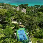Aerial view of the Pickleball Court at NIHI Sumba, surrounded by lush jungle and overlooking the crystal-clear ocean. The vibrant blue court sits harmoniously within the tropical landscape, offering a serene and scenic setting for pickleball enthusiasts.
