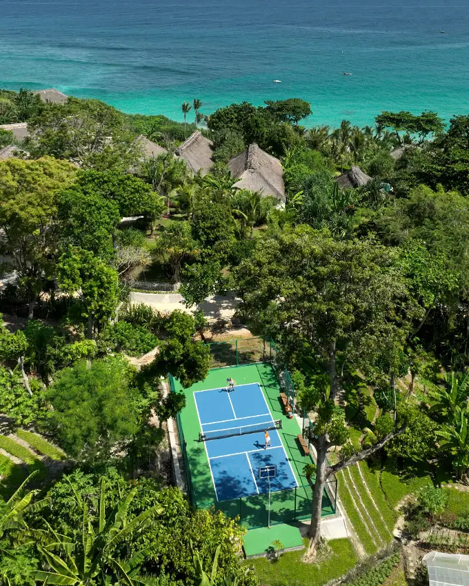 Aerial view of the Pickleball Court at NIHI Sumba, surrounded by lush jungle and overlooking the crystal-clear ocean. The vibrant blue court sits harmoniously within the tropical landscape, offering a serene and scenic setting for pickleball enthusiasts.