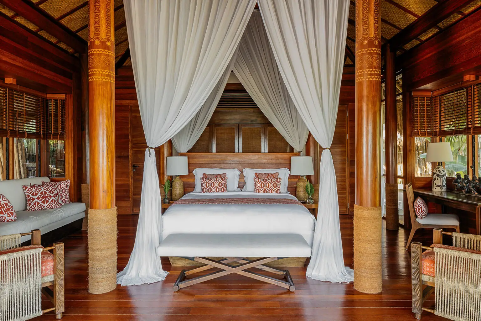 Puncak Sumba House at NIHI® Sumba, featuring a king-size bed with white drapes, surrounded by tropical wooden columns, woven furniture, and soft lighting, creating a serene and luxurious atmosphere.