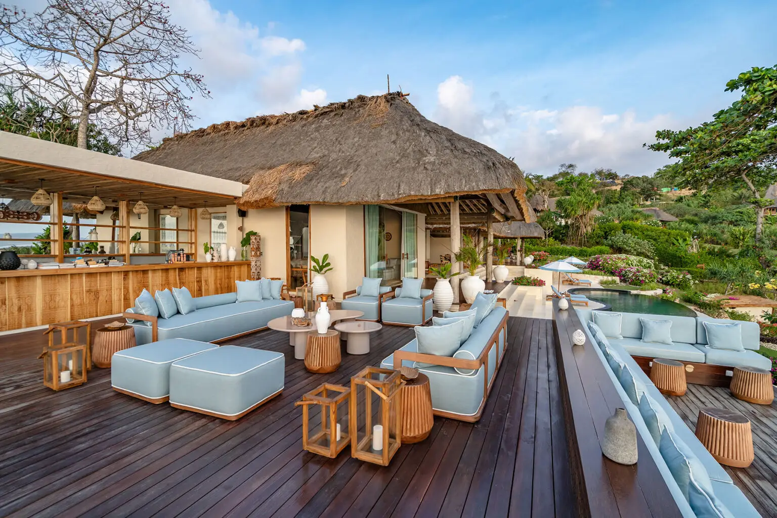 Outdoor lounge area of Putri Kasambi villa, showcasing light blue cushioned seating, thatched roof architecture, a private bar, and a curved pool surrounded by vibrant gardens at NIHI Sumba.