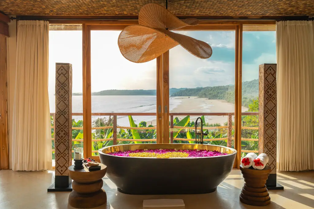Scenic soaking tub filled with flower petals at Putri Kasambi, a two-bedroom villa within NIHI Sumba’s Kasambi Estate, set before floor-to-ceiling glass doors overlooking the serene coastline and lush hills of Sumba.
