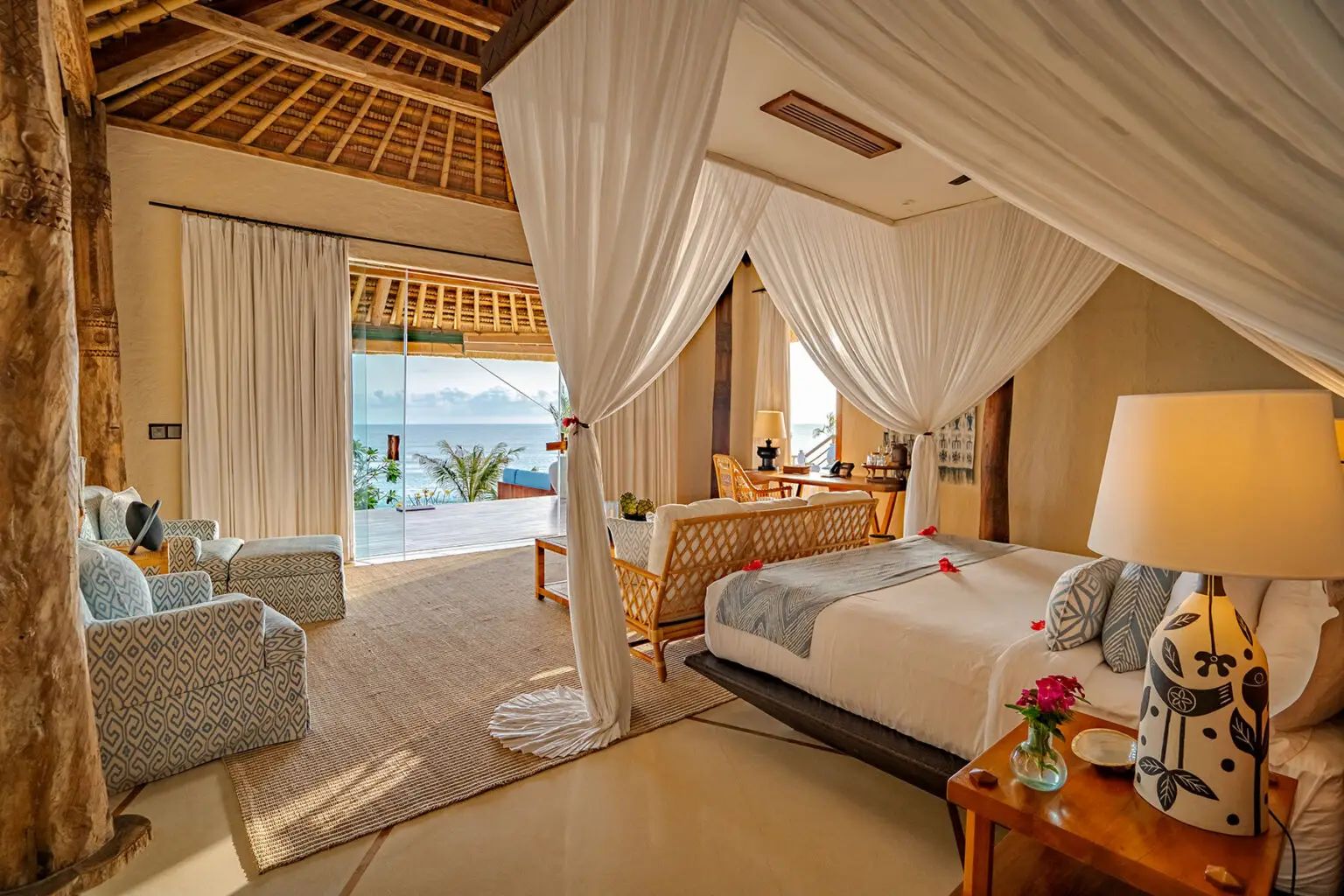 Elegant bedroom at Putri Kasambi villa, featuring a canopy king bed, indoor lounge seating, and sliding glass doors opening to a private terrace with ocean views.