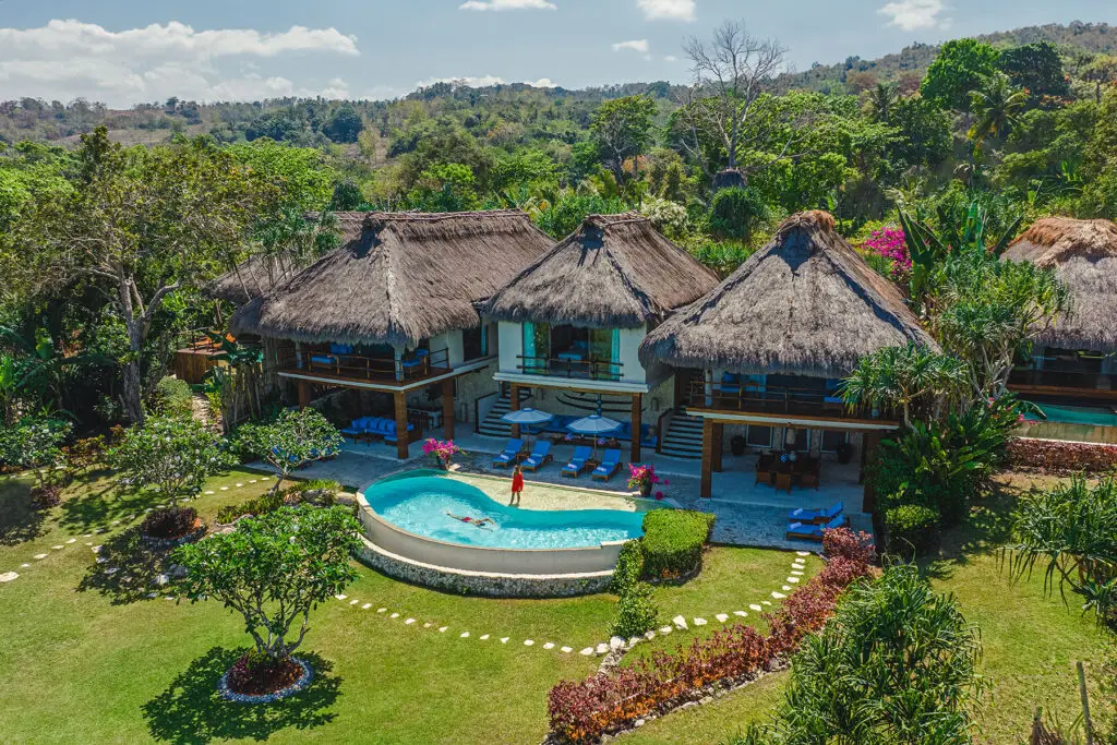 Aerial view of Putri Lantoro Villa at NIHI Sumba, featuring thatched-roof twin-bedroom suites, shaded lounge decks, and a spacious private swimming pool with a guest enjoying a swim, all set against tropical gardens and ocean-facing terraces.
