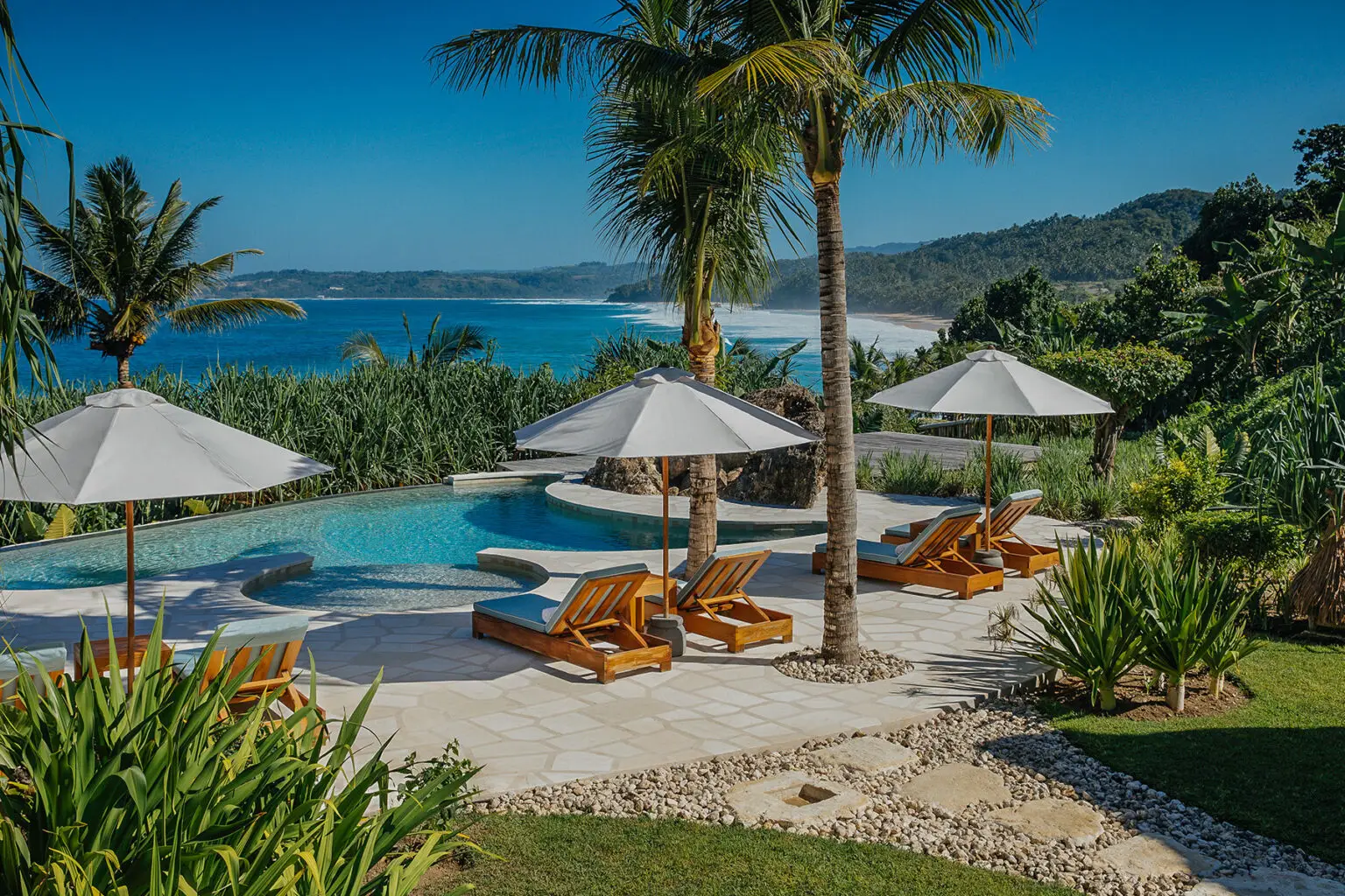 Panoramic view of the private pool area at Putri Wamoro Beach Villa in NIHI Sumba, featuring wooden sun loungers with white umbrellas, swaying palm trees, and a stunning backdrop of the ocean and rugged coastline.