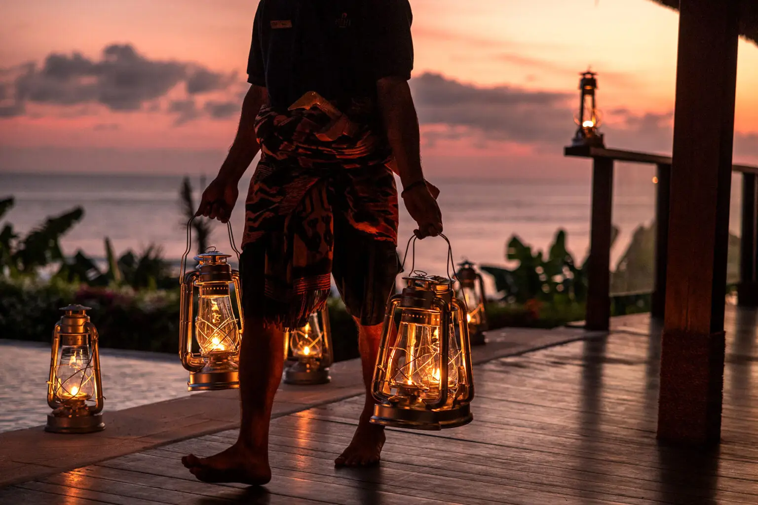 Barefoot NIHI Sumba staff member carrying vintage lanterns at sunset, creating a warm and intimate ambiance overlooking the ocean.